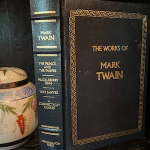 Puede incluir: Un libro de tapa dura azul oscuro titulado The Works of Mark Twain con letras doradas y un borde decorativo. El lomo enumera The Prince and the Pauper, Huckleberry Finn, Tom Sawyer y A Connecticut Yankee, publicado por Longmeadow Press.