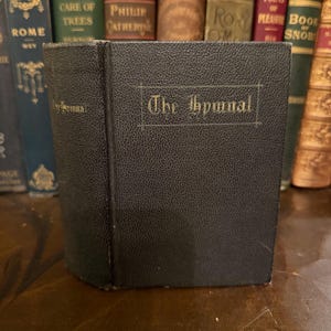 Peut inclure: Un livre gris foncé et texturé intitulé "The Hymnal" avec des lettres dorées. Le livre est debout sur une surface en bois, avec une collection de livres anciens en arrière-plan.