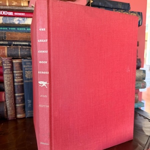 May include: A vintage hardcover book titled "The Great Comic Book Heroes" by Jules Feiffer. The book has a bright red cover and spine with white lettering. The book is part of a collection of antique books.