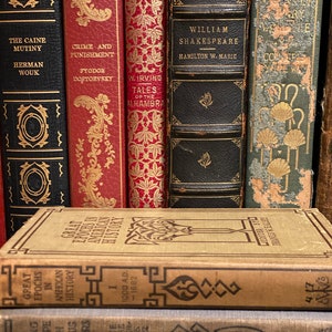 May include: A collection of vintage hardcover books with gold lettering and decorative designs on the spines. The books include titles like "The Caine Mutiny", "Crime and Punishment", "Tales of the Alhambra", "William Shakespeare", and "Great Epochs in American History".