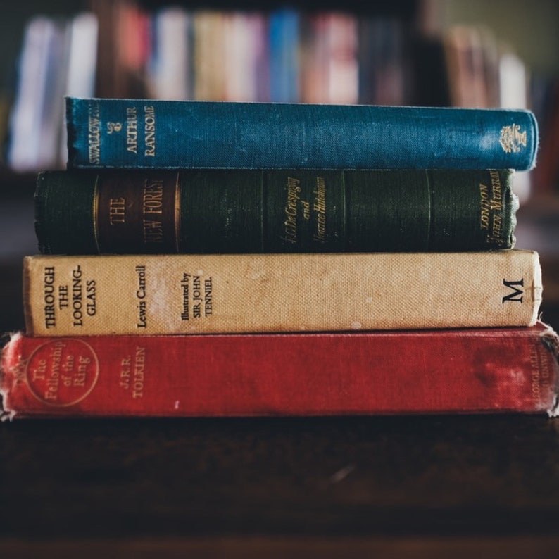 May include: A stack of four hardcover books with colorful spines. The top book is blue with white text, the second book is green with gold text, the third book is cream with brown text, and the bottom book is red with gold text. The text on the spines includes titles like "The New Forest", "Through the Looking Glass", and "The Fellowship of the Ring".