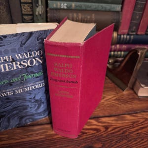 May include: A red hardcover book titled "Ralph Waldo Emerson Essays and Journals" is displayed prominently. The book's spine features gold lettering and a green border. Other books are visible in the background, creating a vintage aesthetic.