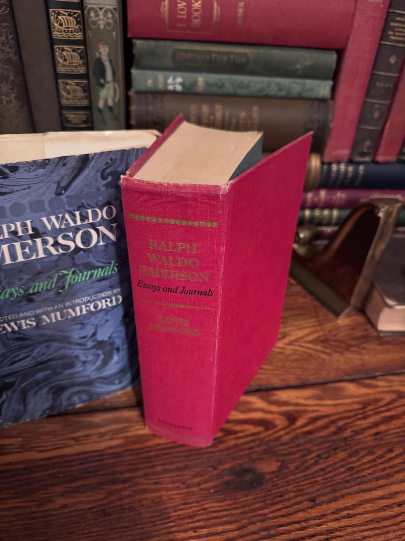 May include: A red hardcover book titled "Ralph Waldo Emerson Essays and Journals" is displayed prominently. The book's spine features gold lettering and a green border. Other books are visible in the background, creating a vintage aesthetic.