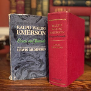 May include: Two vintage books by Ralph Waldo Emerson, "Essays and Journals," are displayed. One book has a dark blue cover with white text, and the other is a red hardcover. The books are on a wooden surface, with other books in the background.