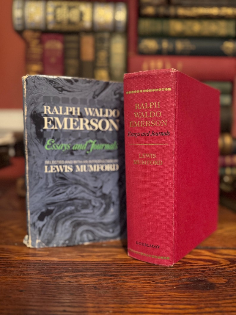 May include: Two vintage books by Ralph Waldo Emerson, "Essays and Journals," are displayed. One book has a dark blue cover with white text, and the other is a red hardcover. The books are on a wooden surface, with other books in the background.
