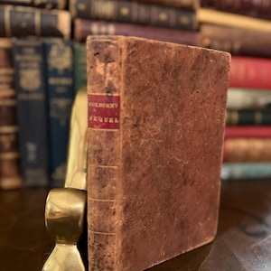 Puede incluir: Un libro antiguo con una cubierta de cuero marrón desgastada. El lomo presenta una etiqueta roja con el título "COLBURN'S SEQUEL". El libro está apoyado por un sujetalibros dorado. Otros libros antiguos son visibles en el fondo.