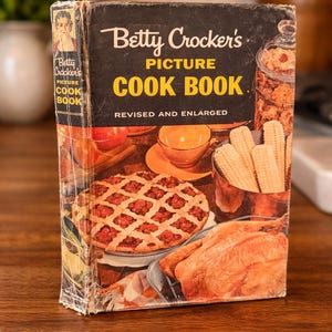 May include: Vintage Betty Crocker's Picture Cook Book. The cover is black with the title in white and yellow. The cover art features a pie, a roasted chicken, and other food items. The book is on a wooden surface.