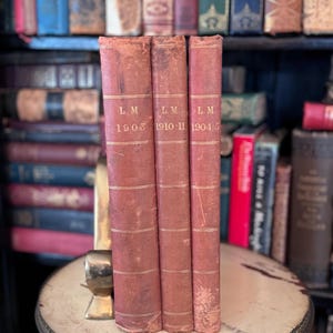 May include: Three antique books with faded red covers and gold lettering, standing upright on a decorative table. The spines display the initials "L.M." and the years 1903, 1910-11, and 1904-5. A collection of other books is visible in the background.