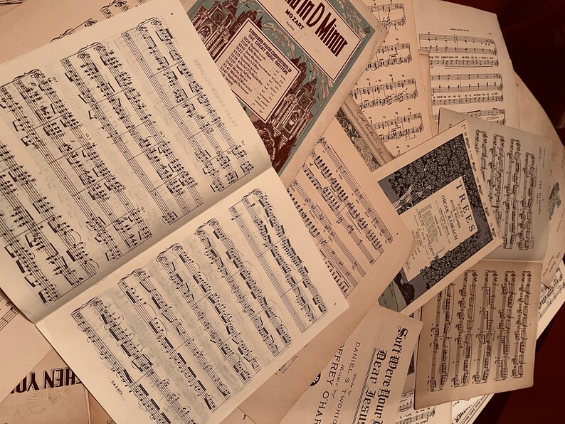 May include: A pile of vintage sheet music with titles like "In D Minor" by Mozart and "Trees" by Oscar Rabach. The music is printed on aged paper with a cream color.