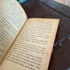 May include: Open book with aged pages, featuring text from a story. The book is open to two pages, with the text visible. The book is resting on a dark surface, with other books visible in the background. The book's pages are yellowed.