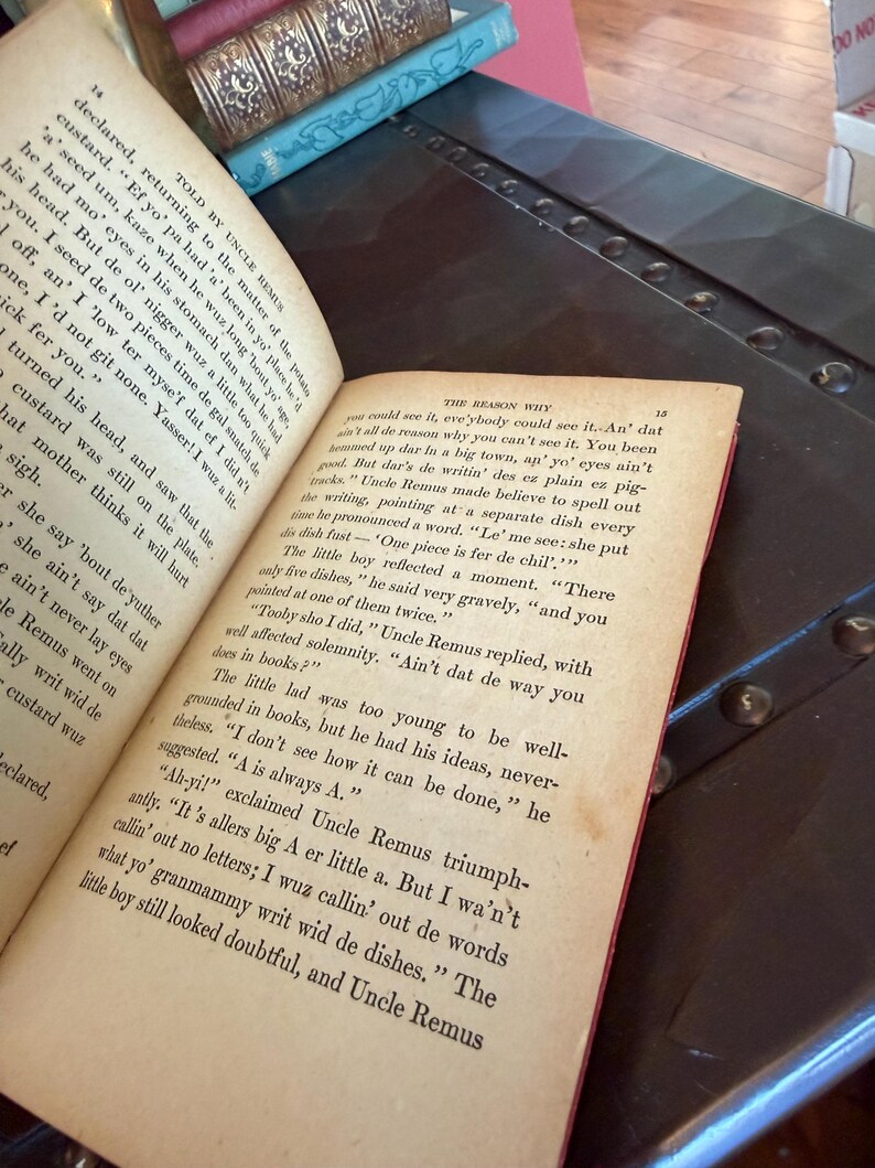 May include: Open book with aged pages, featuring text from a story. The book is open to two pages, with the text visible. The book is resting on a dark surface, with other books visible in the background. The book's pages are yellowed.