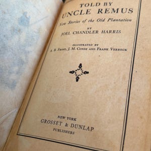 May include: An open book with a tan cover featuring the title "Told by Uncle Remus" and the author's name, Joel Chandler Harris. The book is illustrated by A.B. Frost, J.M. Conde, and Frank Verbeck. Published by Grosset & Dunlap, New York.