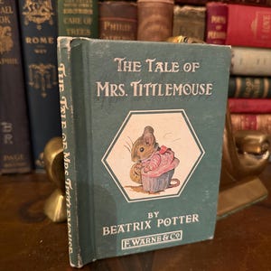 May include: A vintage copy of "The Tale of Mrs. Tittlemouse" by Beatrix Potter. The book has a green cover with the title in white and a color illustration of a mouse. The book is displayed with other antique books in the background.