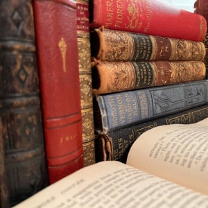 May include: A stack of vintage hardcover books with leather and cloth bindings. The books have gold lettering and decorative designs on the spines. The top book is open and the pages are visible.