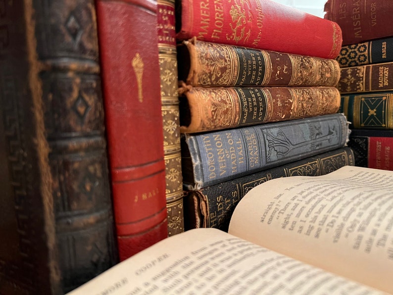 May include: A stack of vintage hardcover books with leather and cloth bindings. The books have gold lettering and decorative designs on the spines. The top book is open and the pages are visible.