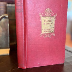 May include: A vintage hardcover book with a red cover and gold lettering. The title "Told by Uncle Remus" is visible, along with the author's name, Joel Chandler Harris. The book is in good condition, with slight wear on the spine and corners.