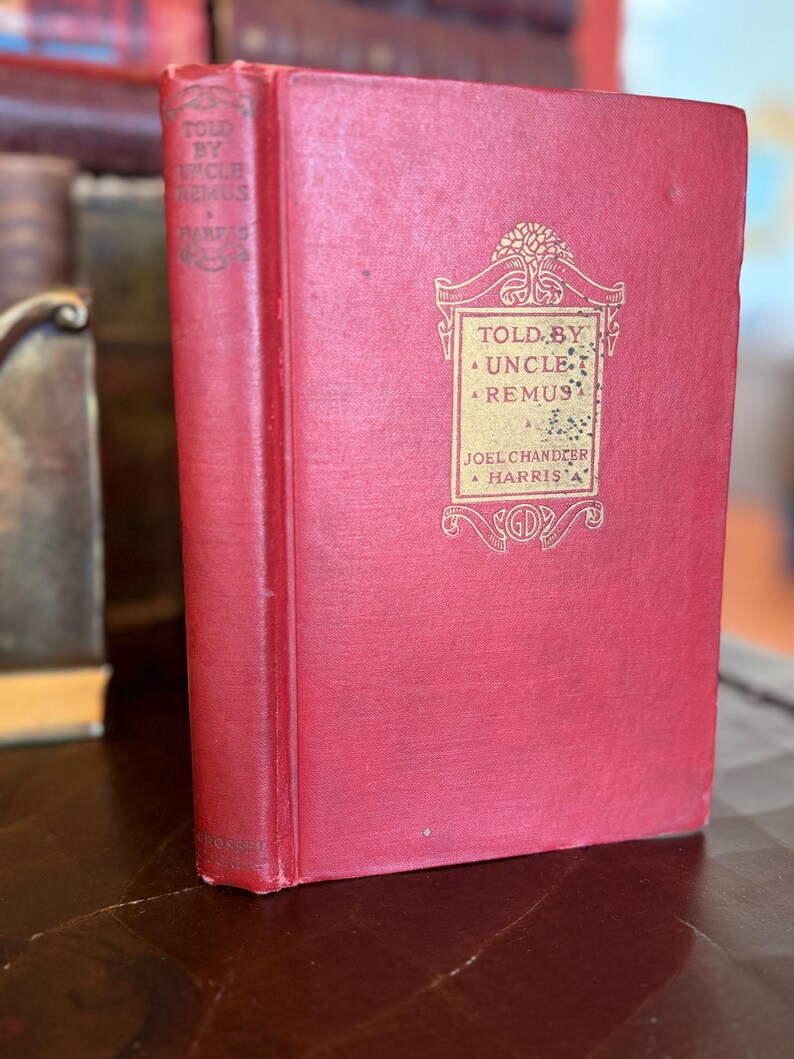 May include: A vintage hardcover book with a red cover and gold lettering. The title "Told by Uncle Remus" is visible, along with the author's name, Joel Chandler Harris. The book is in good condition, with slight wear on the spine and corners.