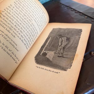 May include: An open antique book with aged, yellowed pages and red binding. A black and white illustration of a person is visible on the right page, with the text "So he holler down thoo de crack".
