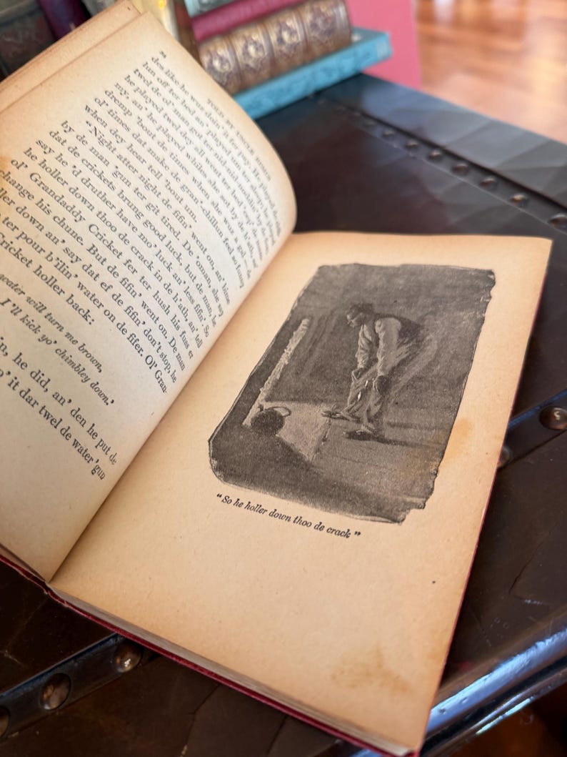 May include: An open antique book with aged, yellowed pages and red binding. A black and white illustration of a person is visible on the right page, with the text "So he holler down thoo de crack".