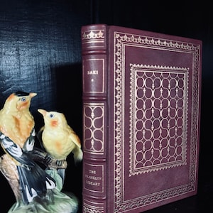 Può includere: Un libro rilegato in pelle bordeaux con dettagli dorati e il titolo "SAKI" sul dorso. Il libro fa parte della collezione "The Franklin Library". Due statuette di uccelli in ceramica sono in primo piano.