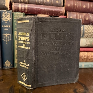 May include: A black, vintage book titled "Audels Pumps Hydraulics Air Compressors" with gold lettering. The book is part of a collection of antique books, with the spine visible, and the text "A Practical Treatise".