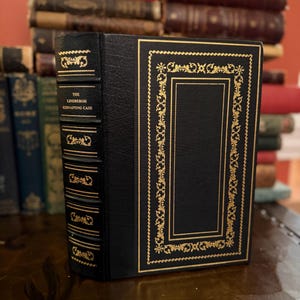 Könnte beinhalten: Ein schwarzes Hardcover-Buch mit dem Titel "The Lindbergh Kidnapping Case" und goldenen Verzierungen. Das Buch steht aufrecht und zeigt seinen Buchrücken und die Vorderseite. Im Hintergrund sind weitere Bücher zu sehen, was auf eine Bibliothek hindeutet.
