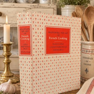 May include: A hardcover book titled Mastering the Art of French Cooking by Julia Child, Louisette Bertholle, and Simone Beck. The cover features a cream background with a repeating red and blue fleur-de-lis pattern and red rectangular text boxes.