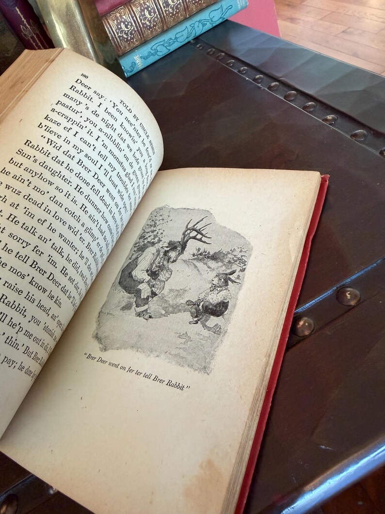 May include: An open vintage book with a red spine, displaying an illustration of two anthropomorphic animals. The text reads "Brer Deer wend on jer ter tell Brer Rabbit". Other books are visible in the background.
