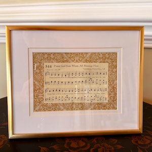Puede incluir: Obra de arte enmarcada con partituras titulada "Praise God from Whom All Blessings Flow". El marco dorado rodea un paspartú blanco y una impresión de estilo vintage con notas musicales y texto. Un elemento decorativo.