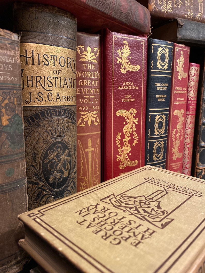 May include: A close-up of a bookshelf with a variety of antique books. The books have leather covers and gold lettering. The titles of the books include "History of Christianty", "The World's Great Events", "Anna Karenina", and "The Caine Mutiny".