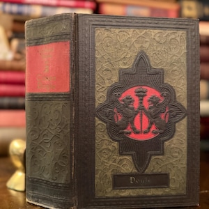 Op de afbeelding: Antiek boek met een rode rug en een decoratieve omslag. De omslag heeft een centraal embleem met twee gevleugelde wezens en een kroon, tegen een groene en bruine achtergrond. Het woord "Doub" is zichtbaar op de omslag.