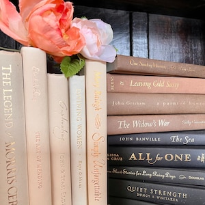 Puede incluir: Una colección de libros de tapa dura en varios tonos de beige, crema y marrón, colocados en una estantería. Letras doradas adornan los lomos, con títulos como "Simply Unforgettable" y "The Sea". Un arreglo floral color melocotón descansa sobre los libros.
