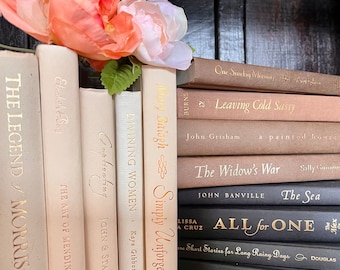 May include: A collection of hardcover books in various shades of beige, cream, and brown, arranged on a shelf. Gold lettering adorns the spines, with titles like "Simply Unforgettable" and "The Sea." A peach-colored floral arrangement rests atop the books.