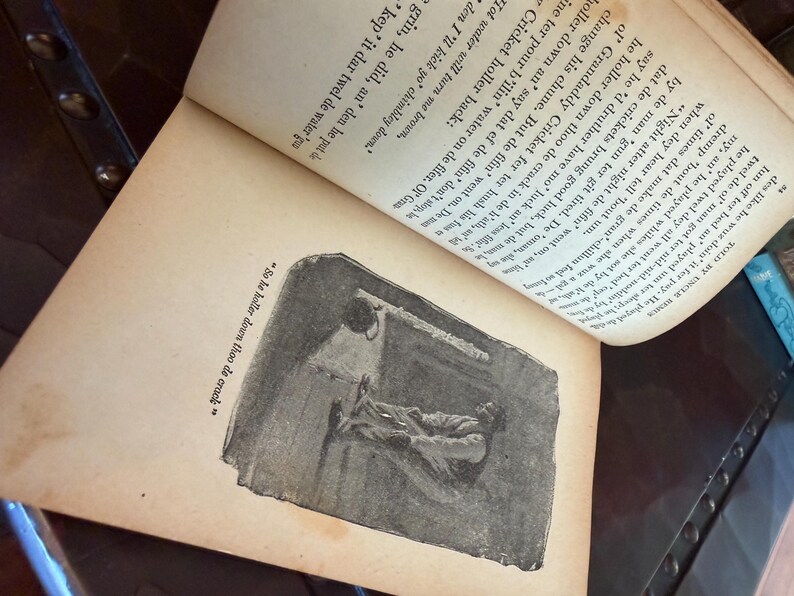 May include: An open book with aged, yellowed pages. The text is in black ink, with an illustration of a person lying down. The illustration is in black and white. The text reads "So he holler down thoo de crack."