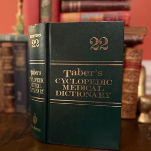 Puede incluir: Un libro verde oscuro "Taber's Cyclopedic Medical Dictionary", edición 22, con letras doradas. El libro está de pie, con otros libros en el fondo.