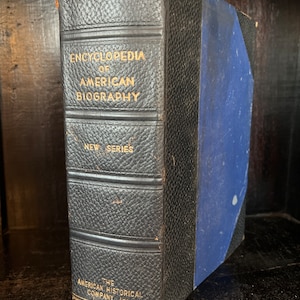 May include: A black leather-bound book titled "Encyclopedia of American Biography, New Series" with gold lettering. The book is published by The American Historical Company, Inc.