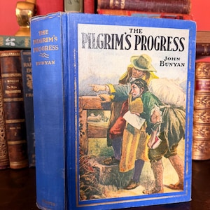 May include: A vintage copy of "The Pilgrim's Progress" by John Bunyan. The book has a blue cover with gold lettering and a color illustration. The cover art depicts two men in period clothing. The book is surrounded by other antique books.