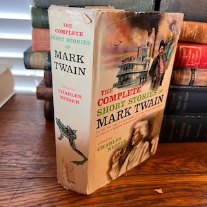 Op de afbeelding: Een hardcover boek getiteld The Complete Short Stories of Mark Twain, onder redactie van Charles Neider. De crèmekleurige stofomslag bevat een illustratie van een groene kikker op de rug en een afbeelding van een stoomboot en Mark Twain.