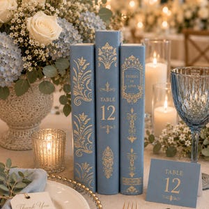 May include: Three vertical dusty blue books with gold foil filigree and text, including the centre book labelled Table 12 and the right book labelled Stories of Love, accompanied by a matching blue Table 12 card and a Thank You for celebrating with us tag.