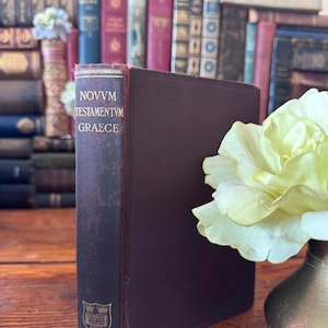 May include: A maroon-colored book with gold lettering, titled "NOVVM TESTAMENTVM GRAECE" and the word "OXFORD" at the bottom. The book is standing upright in front of a blurred background of other books. A yellow flower is in a small vase.