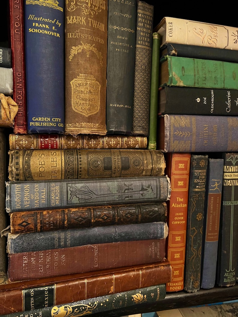 May include: A bookshelf filled with vintage hardcover books. The books are arranged in stacks and have various colors, including brown, blue, green, and red. Some of the books have titles visible, such as "Mark Twain Illustrated", "The Alaskan", and "St. George for England".
