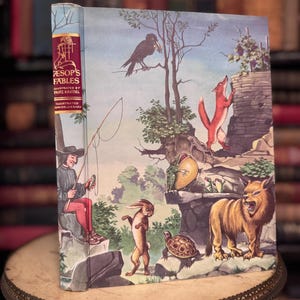 Könnte beinhalten: Ein Vintage-Exemplar von Äsops Fabeln, illustriert von Fritz Kredel. Das Cover zeigt eine farbenfrohe Illustration von Tieren und einem Mann beim Angeln. Das Buch wird auf einem dekorativen Ständer präsentiert.
