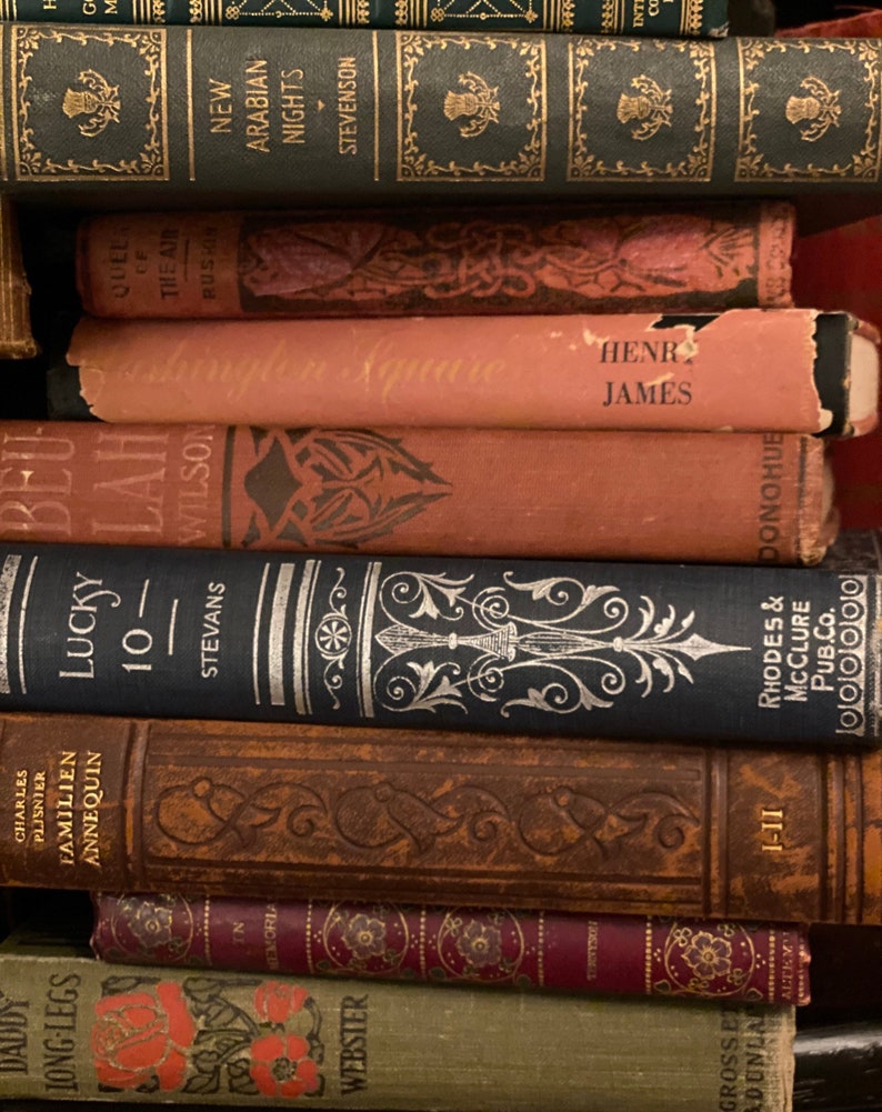 May include: A stack of vintage hardcover books with various titles and authors. The books are arranged in a vertical stack, with the top book being a red hardcover with the title "Henry James". The books are all in good condition and have a worn, aged look.