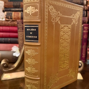 以下が含まれることがあります： 「Molière Comédies」というタイトルのヴィンテージハードカバー本。茶色の革の表紙と金色のエンボス加工の装飾が施されています。背表紙には黒い文字でタイトルが記されています。本は直立して表示され、その華やかなデザインが際立っています。