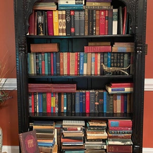 May include: A black wooden bookcase with ornate carvings, filled with books. The bookcase has five shelves and is decorated with a gold metal pheasant figurine. The books are arranged in a variety of sizes and colors, and some are stacked on top of each other. There are also a few books on the floor in front of the bookcase.