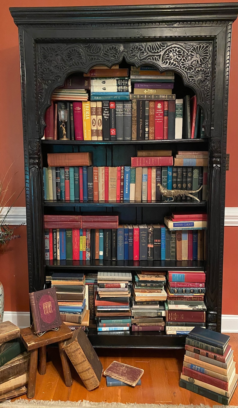 May include: A black wooden bookcase with ornate carvings, filled with books. The bookcase has five shelves and is decorated with a gold metal pheasant figurine. The books are arranged in a variety of sizes and colors, and some are stacked on top of each other. There are also a few books on the floor in front of the bookcase.