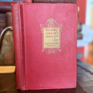 May include: A vintage hardcover book titled "Told by Uncle Remus" by Joel Chandler Harris. The book has a red cover with gold lettering and a decorative emblem. The spine is also red with the title visible.