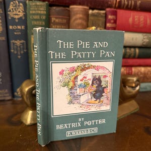 Peut inclure: Un exemplaire vintage de "The Pie and The Patty Pan" de Beatrix Potter. Le livre a une couverture turquoise avec des lettres blanches et une illustration d'un chat noir. Il est présenté devant une collection de livres anciens.
