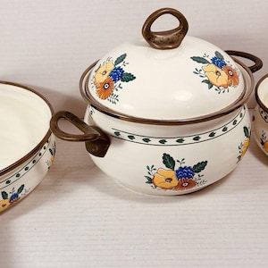 Puede incluir: Un juego de tres piezas de cocina de esmalte color crema con bordes y asas de color marrón. El conjunto incluye una sartén poco profunda, una olla con tapa y un cazo, decorados con un patrón floral de flores amarillas, naranjas y azules.