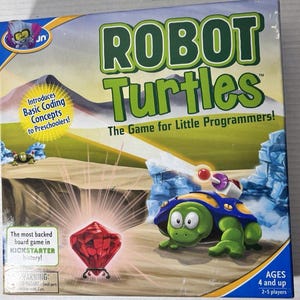 Puede incluir: Una caja de juego de mesa Robot Turtles con una tortuga verde de caparazón azul y una gema roja sobre un soporte. El texto dice Robot Turtles, The Game for Little Programmers, Introduces Basic Coding Concepts to Preschoolers y The most backed board game in KICKSTARTER history.